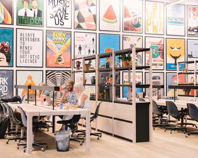 Two people sit at a long table in a bright, art-filled communal workspace.