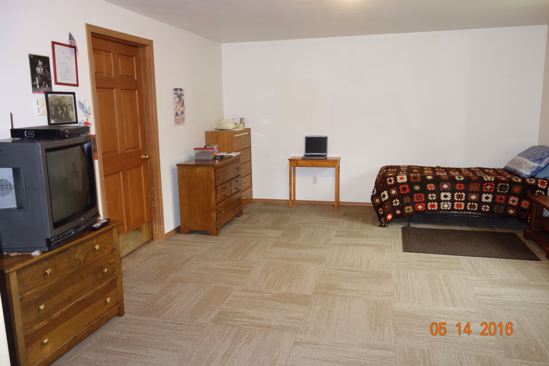 A simple room with beige carpet tiles, a wooden door, a wooden dresser with a TV on top, another wooden dresser, a small wooden table with a laptop, and a single bed with a colorful crocheted blanket and pillows.