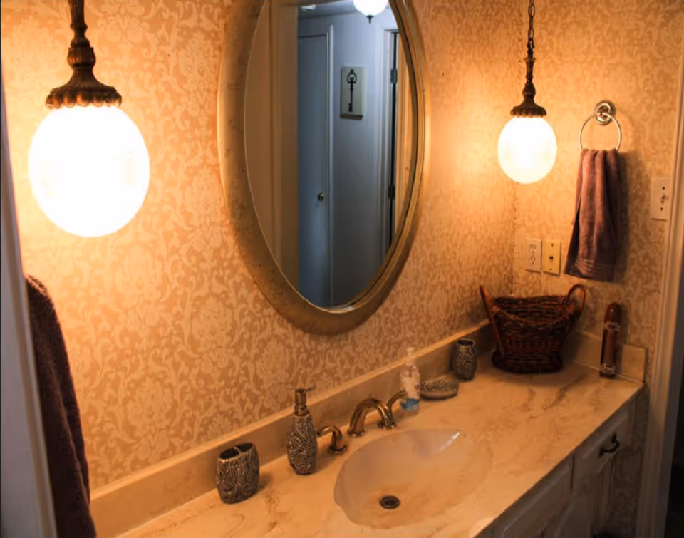 Elegant bathroom vanity with oval mirror, warm wallpaper, towels, and countertop essentials.