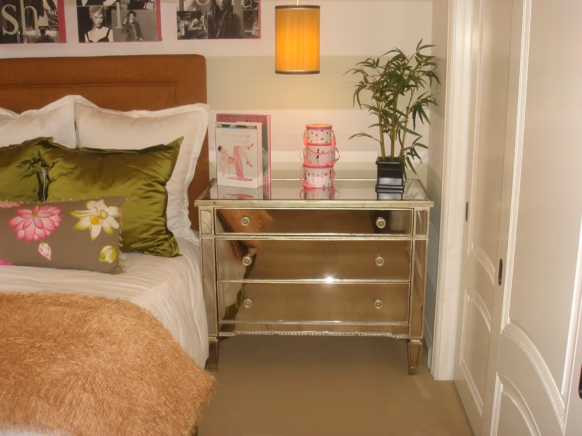 Bed with green and floral pillows next to a mirrored nightstand topped with a hanging lamp and potted plant.