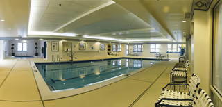 Indoor swimming pool area with clear water, surrounded by beige tiled flooring. Several white and black lounge chairs are lined up along the right side. The ceiling has recessed lighting and decorative molding. Windows and framed artwork are visible on the far walls.