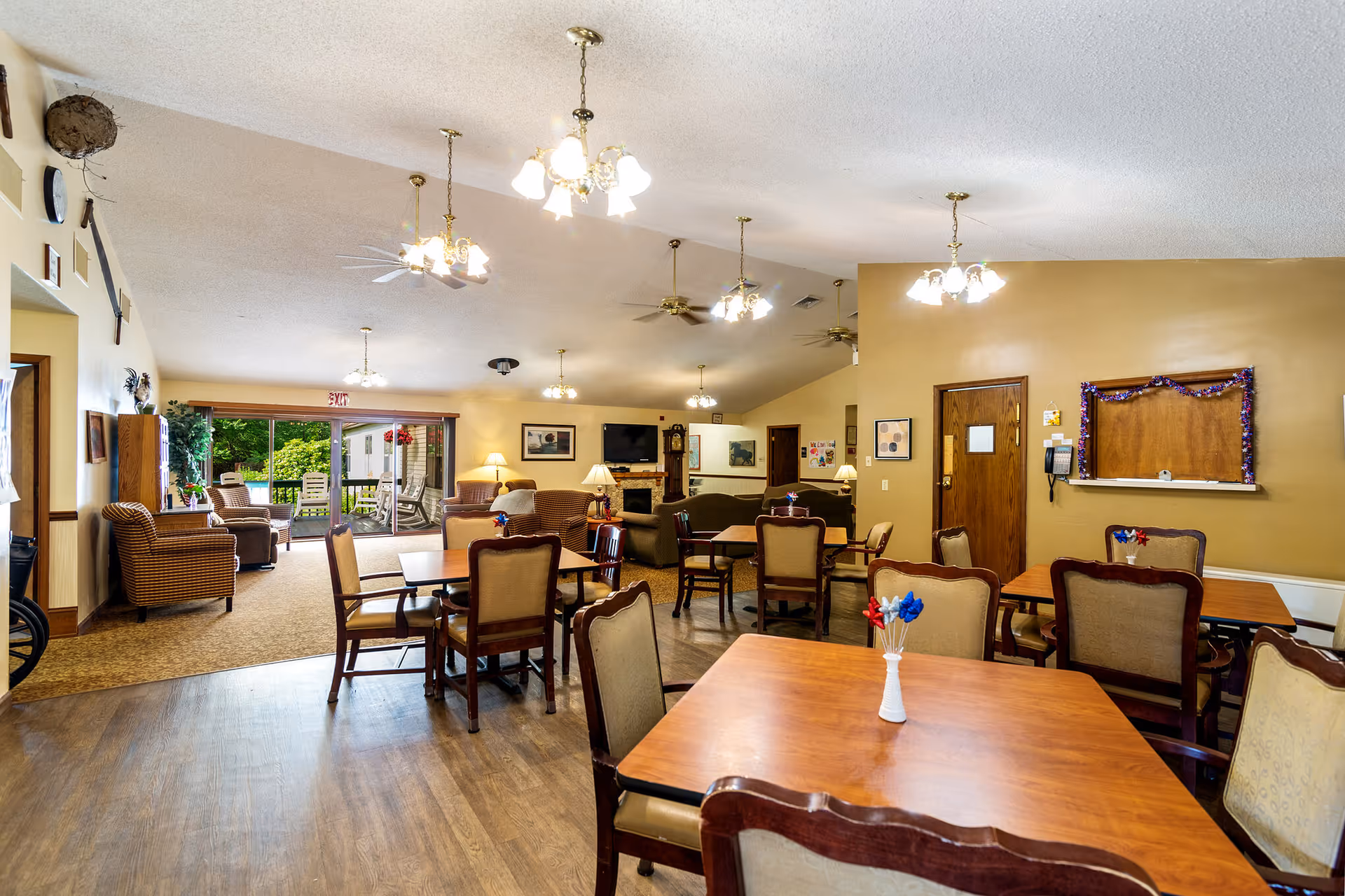 A spacious senior living common area with multiple wooden tables and cushioned chairs arranged for dining or socializing. The room features warm beige walls, several ceiling lights, ceiling fans, and a carpeted seating area with armchairs near large sliding glass doors that open to an outdoor patio with greenery. A fireplace and a wall-mounted TV are visible in the background.