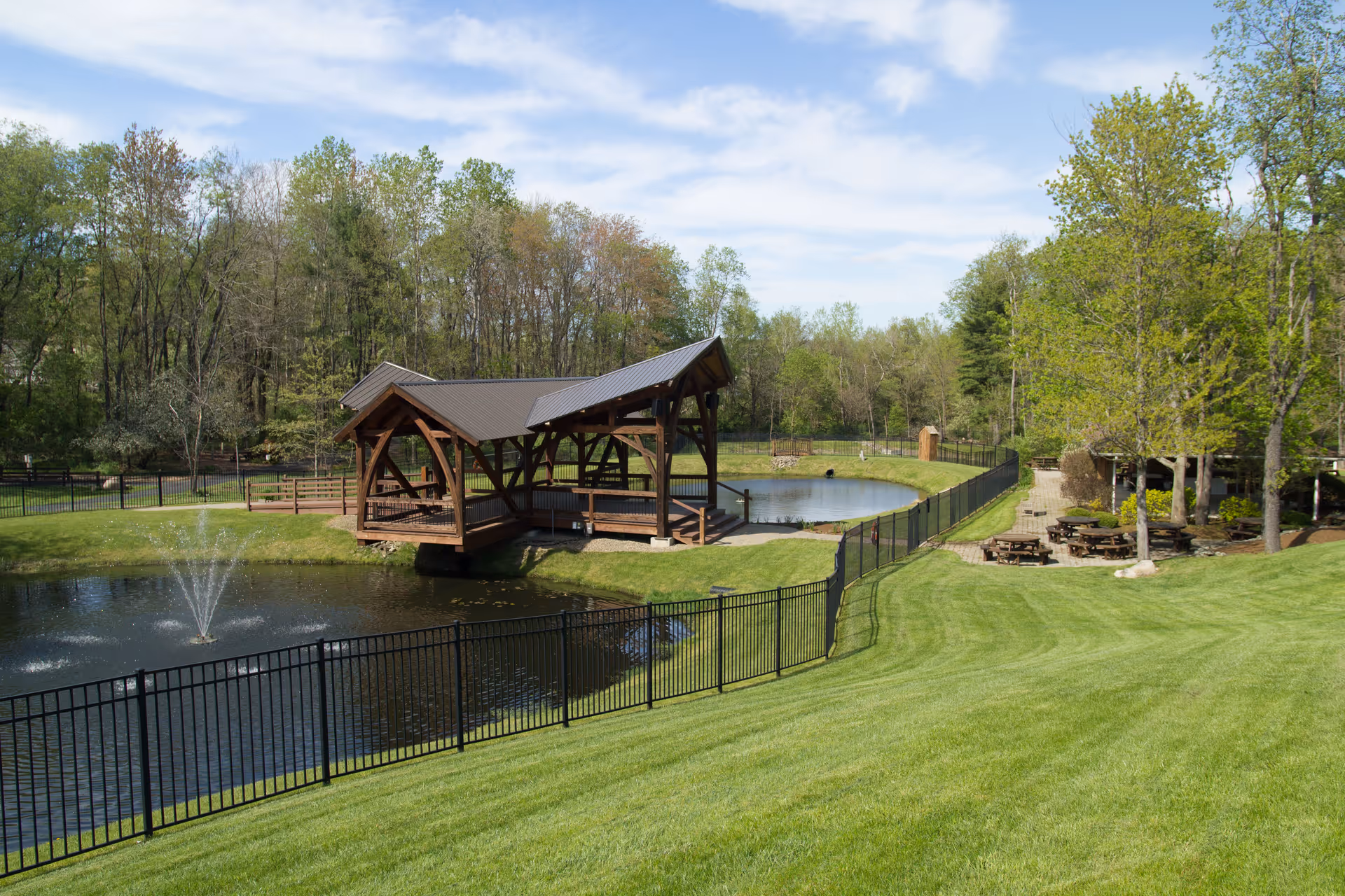 A serene outdoor area at Memory Garden featuring a small pond with a water fountain, a covered wooden gazebo bridge over the pond, surrounded by green grass and trees. Picnic tables are visible near the trees on the right side under a clear blue sky with some clouds.
