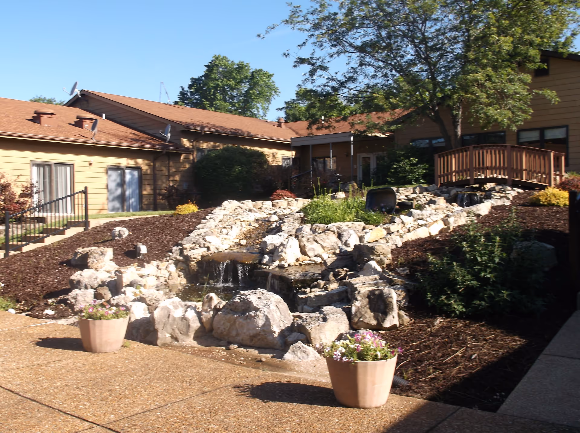 Outdoor courtyard with a rock water feature, small wooden bridge, potted flowers, and a low building in the background.