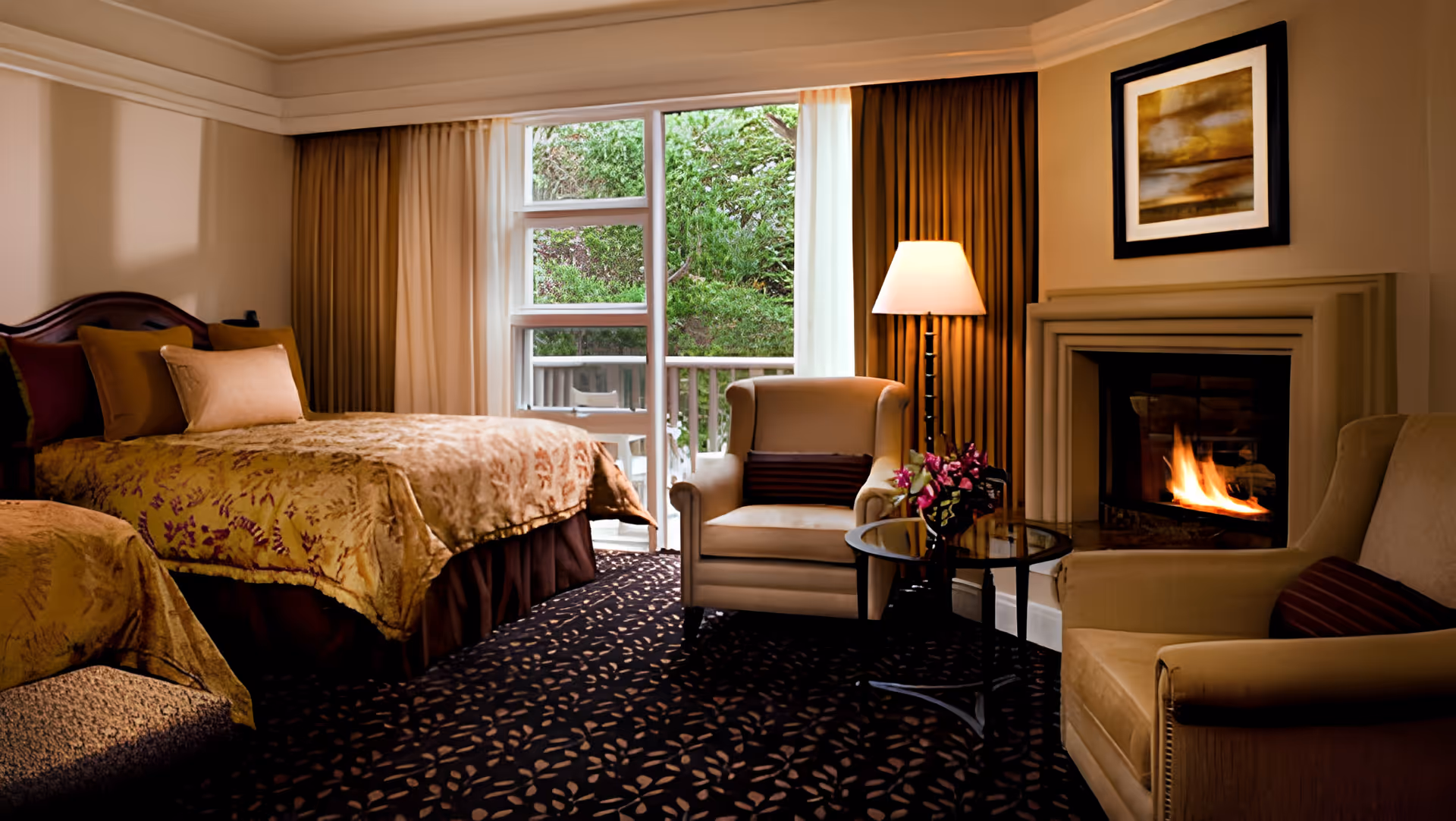 A cozy bedroom with two beds covered in golden bedspreads, a pair of beige armchairs with a small round glass table between them, a lit fireplace, a floor lamp, and a large window with curtains showing green foliage outside.