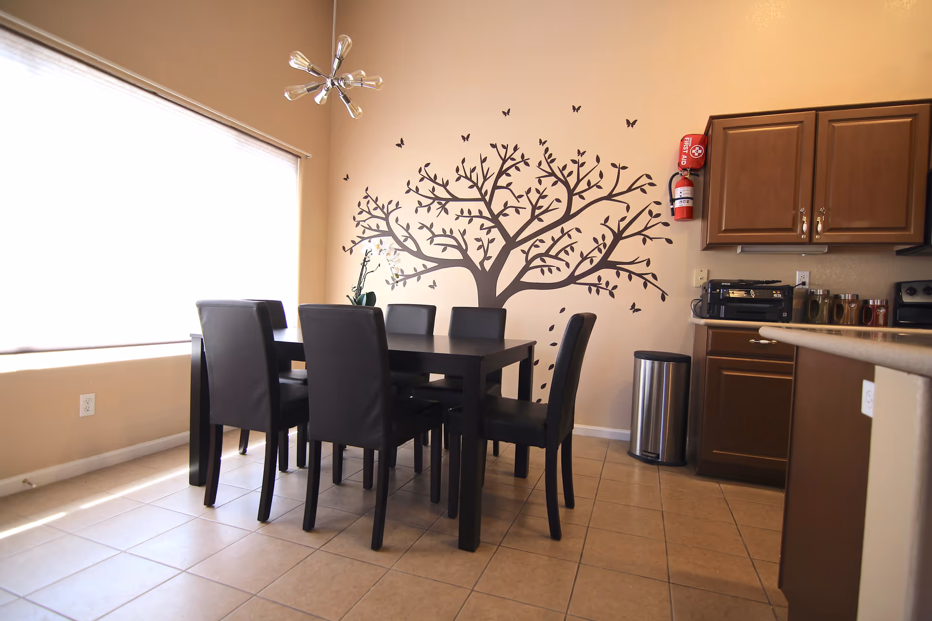 A dining area with a black dining table and six black chairs on a tiled floor. The wall behind the table features a large tree and butterfly decal. To the right, there is a kitchen counter with brown cabinets, a fire extinguisher mounted on the wall, a trash can, and some kitchen appliances. A large window with a white blind is on the left side, allowing natural light to fill the room.