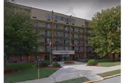 Multi-story senior living building with balconies, trees, and a walkway to the entrance.