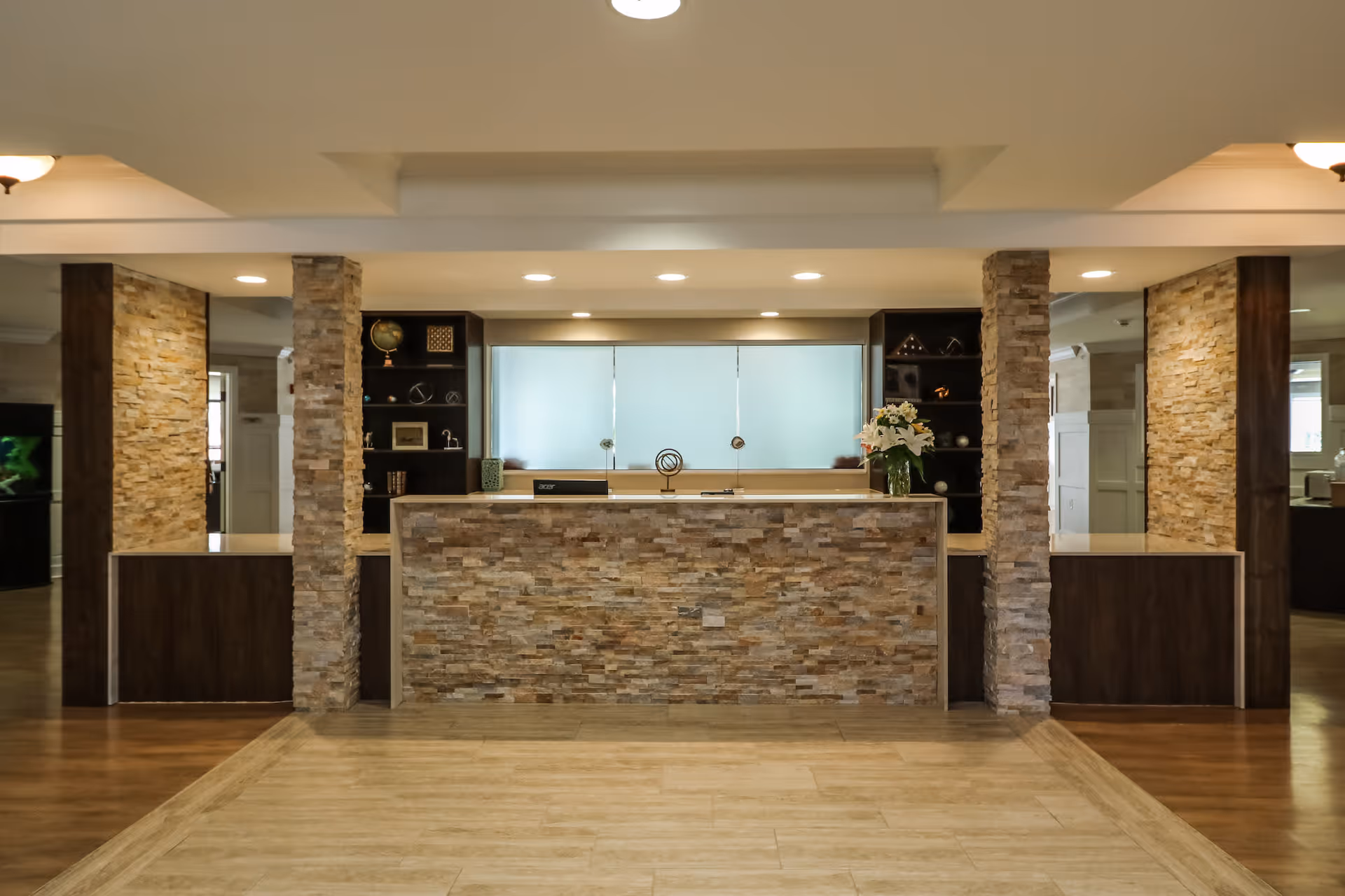 Reception desk area with a stone facade and wooden accents, flanked by stone pillars and shelves with decorative items, under recessed ceiling lights.