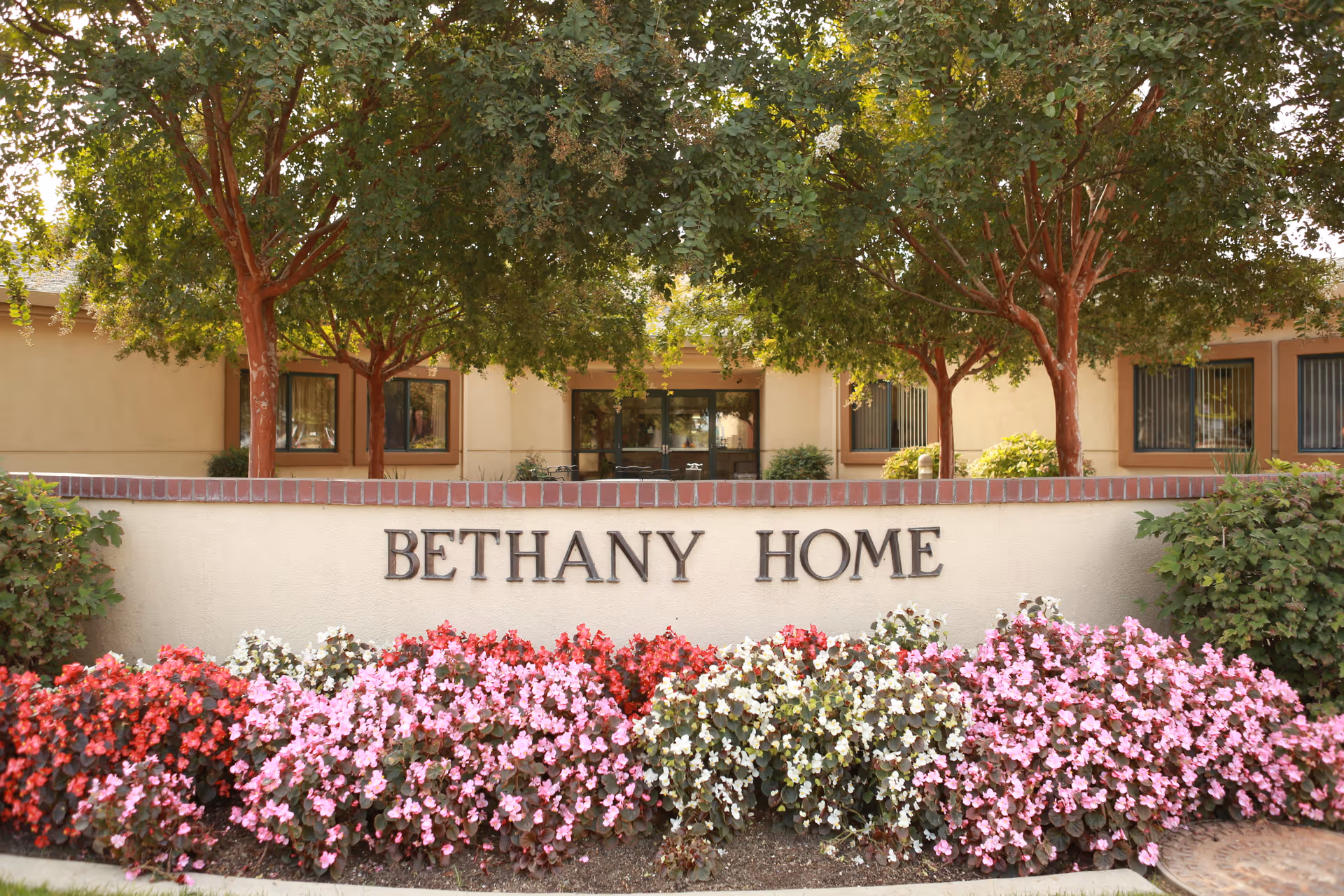 Front entrance sign reading "Bethany Home" with colorful flower beds and trees in front of the building.