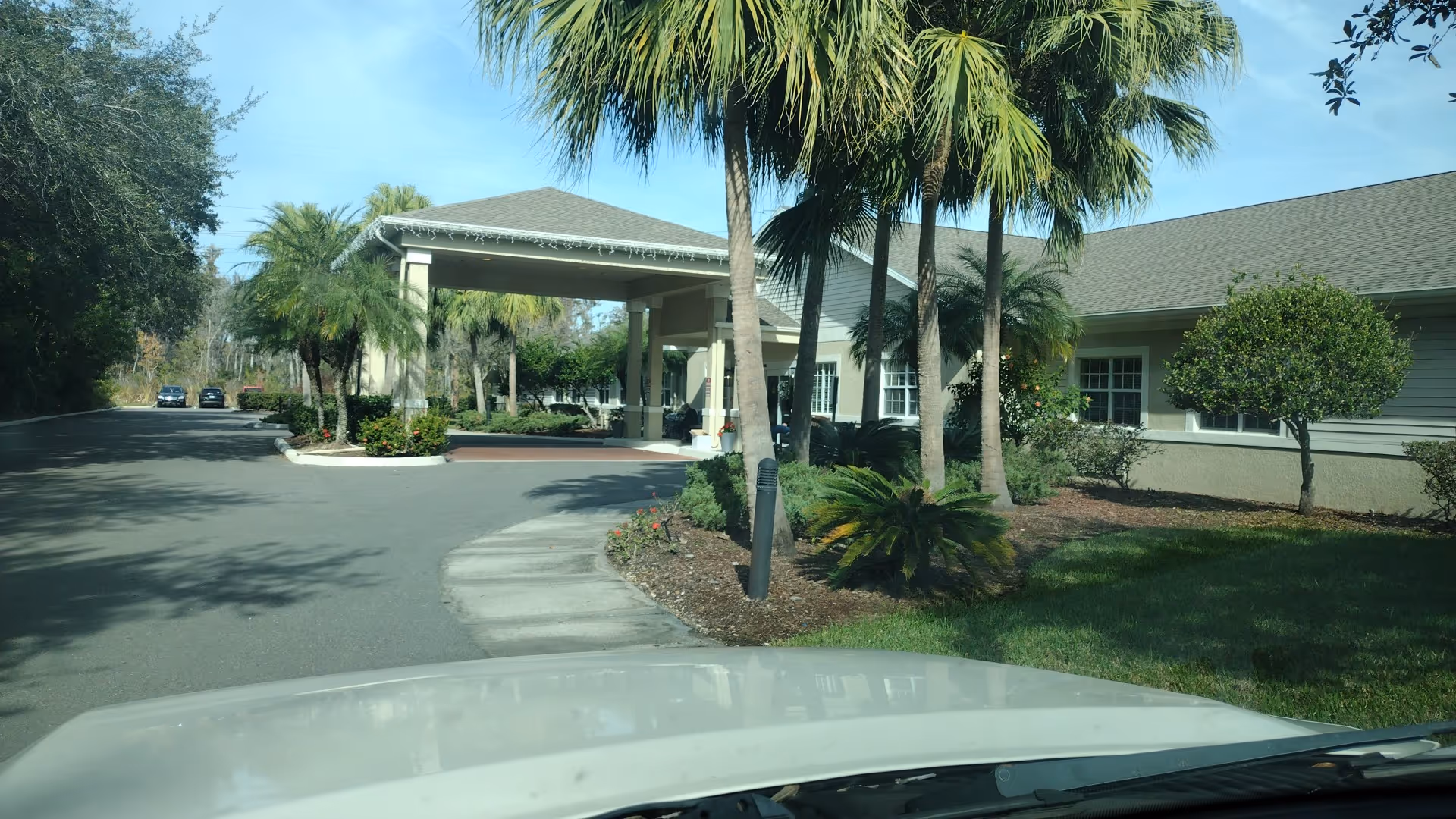 Entrance to Aston Gardens the Inn adult living facility with a curved driveway and palm trees.