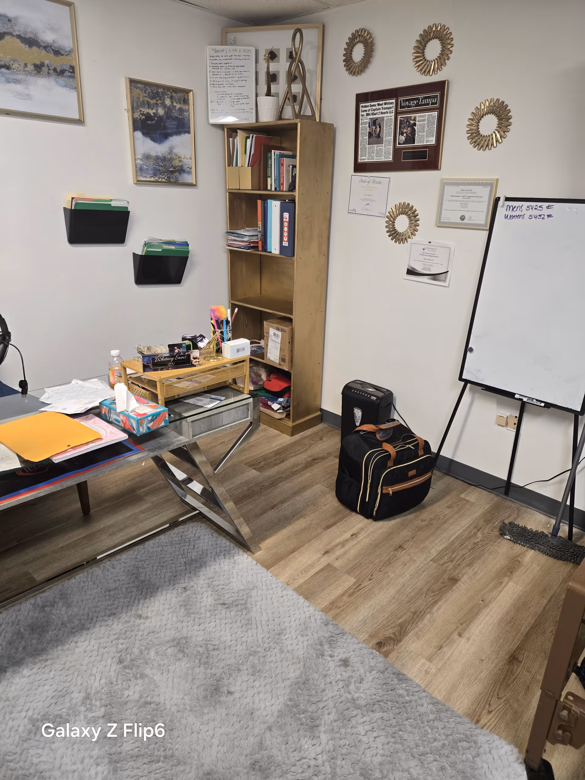 Office-like room with desk, bookshelf, whiteboard, and wall art.