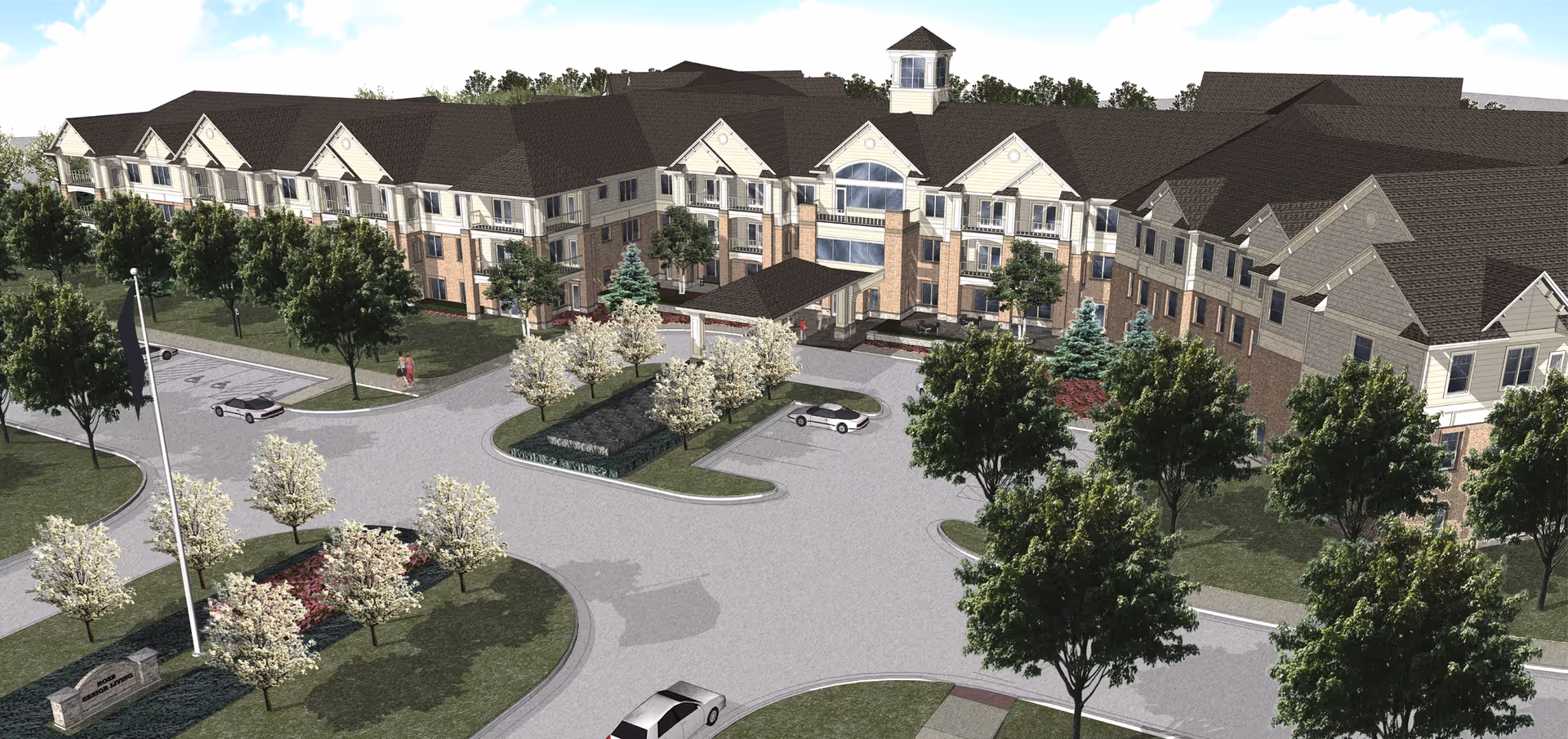Aerial view of a large senior living facility building with multiple floors, surrounded by trees and landscaped grounds. There is a circular driveway with parked cars and a flagpole near the entrance. The building has a combination of brick and light-colored siding with a dark roof.