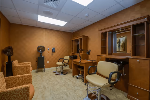 Small salon-style room with styling chairs, hair dryers, wooden cabinets and workstations under fluorescent ceiling lights.