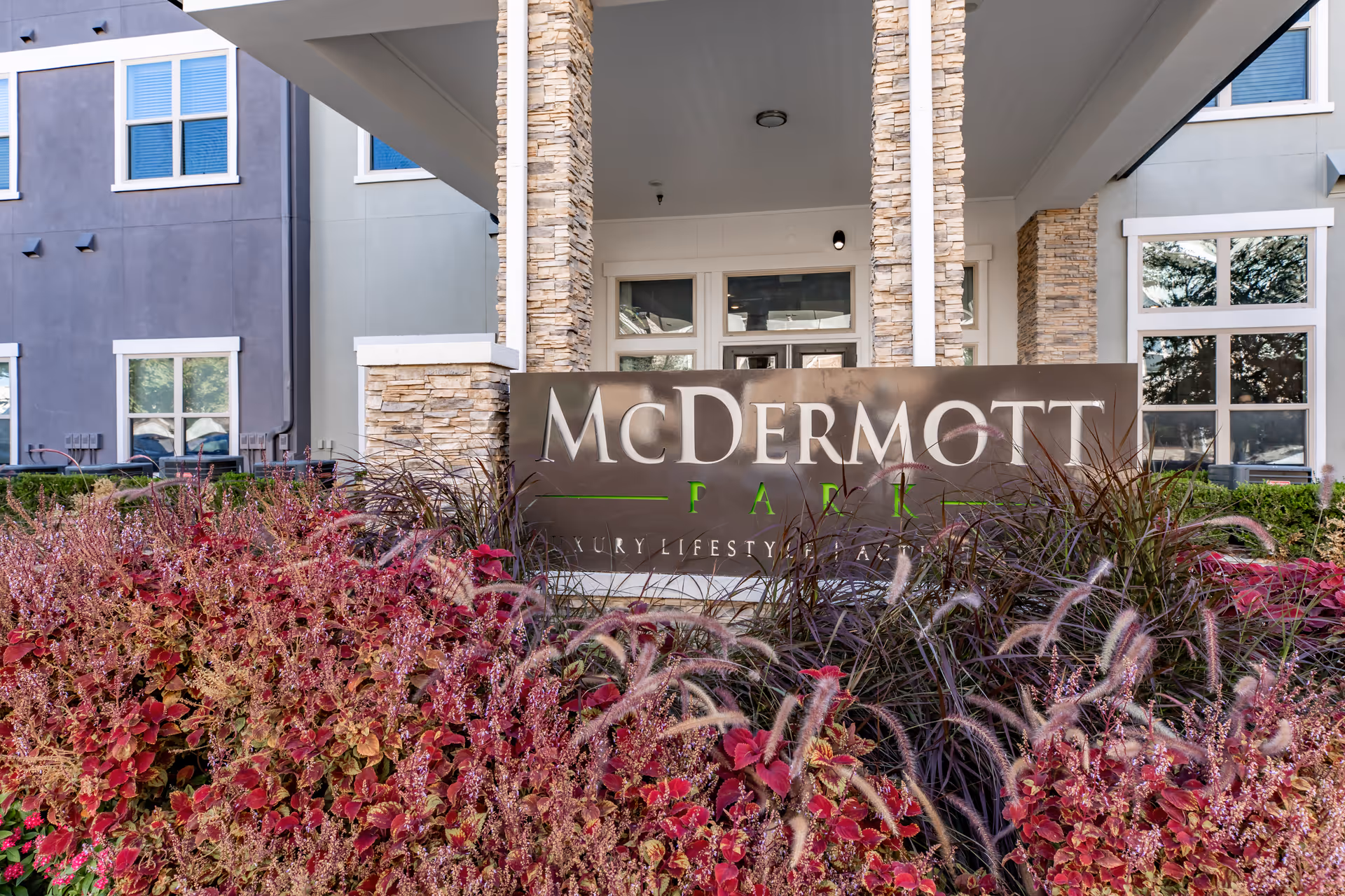 Stone-pillared entrance with a sign reading "McDermott" surrounded by ornamental grasses and red foliage in front of the building.