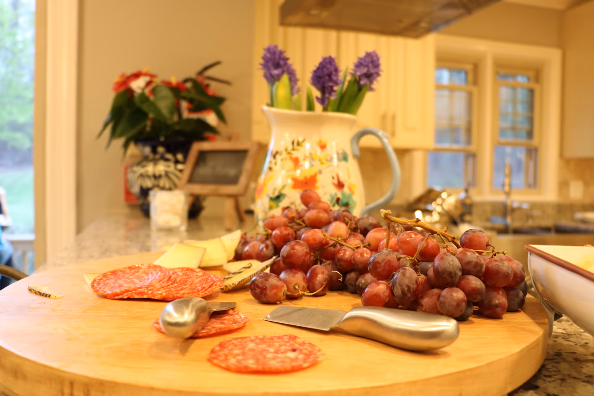 Cutting board with grapes, sliced salami and cheese on a kitchen island with a floral pitcher and cabinets in the background.