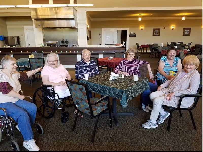 A group of six elderly people, including two in wheelchairs, sitting around a table with a green patterned tablecloth in a spacious dining area. The background shows more tables and chairs, a kitchen area with stainless steel appliances, and warm lighting on the walls.