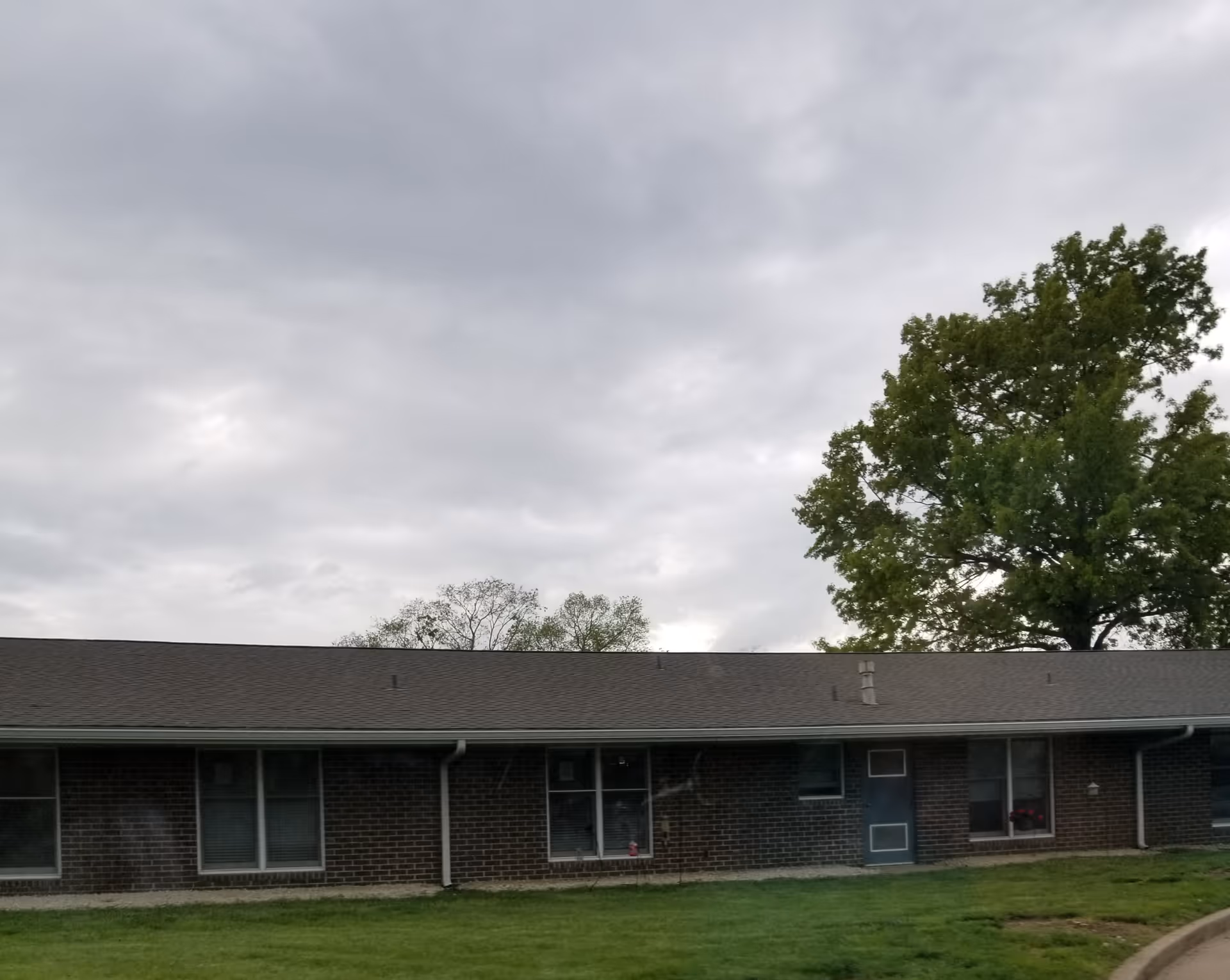 One-story brick senior living facility exterior with windows, lawn, and a large tree.