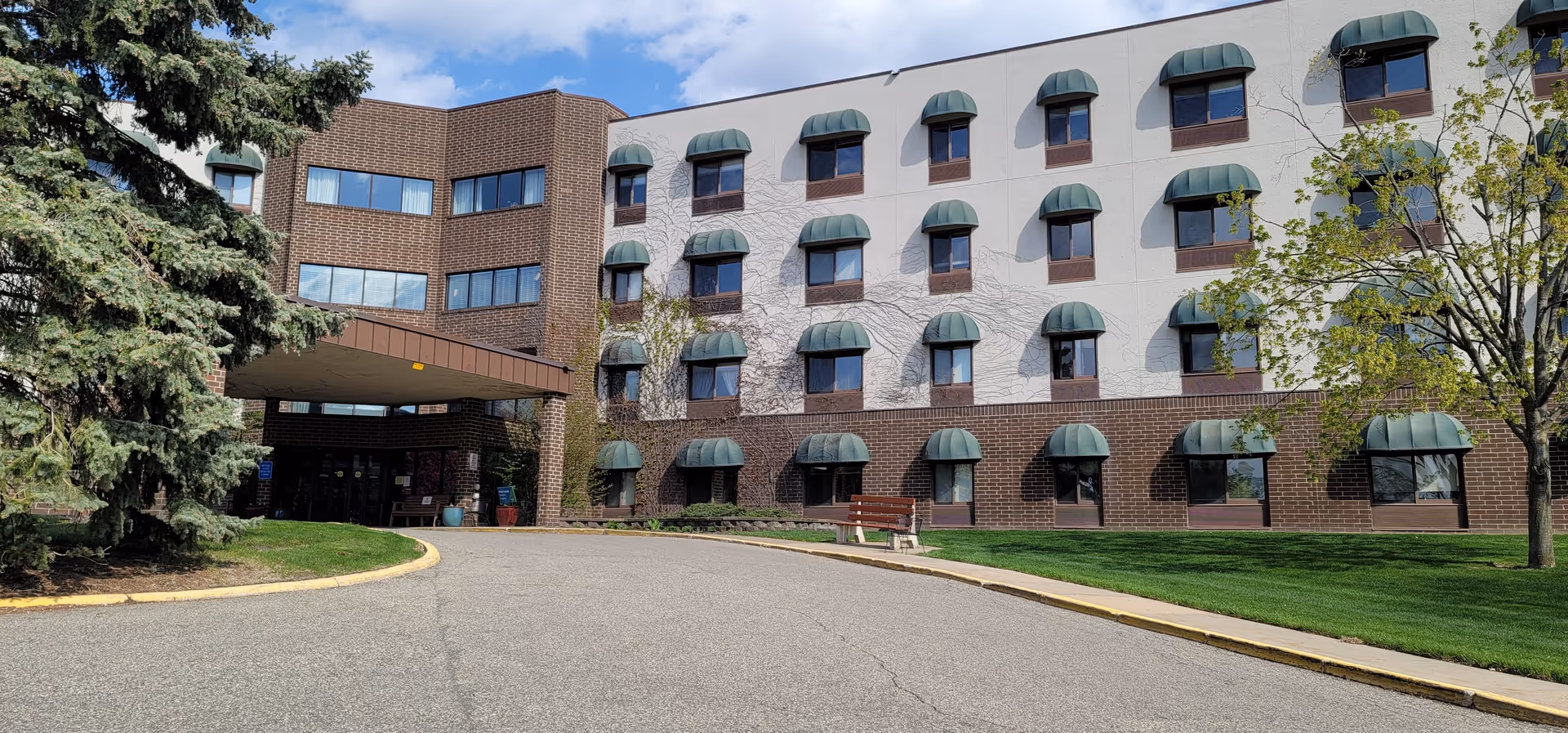 Exterior view of a multi-story senior living facility building with brown brick and white walls, featuring multiple windows with green awnings. There is a covered entrance with a driveway leading up to it, surrounded by green grass, trees, and a bench near the entrance.