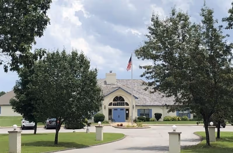 Front entrance of a single-story senior living building with a flagpole, circular driveway, and trees on the lawn.
