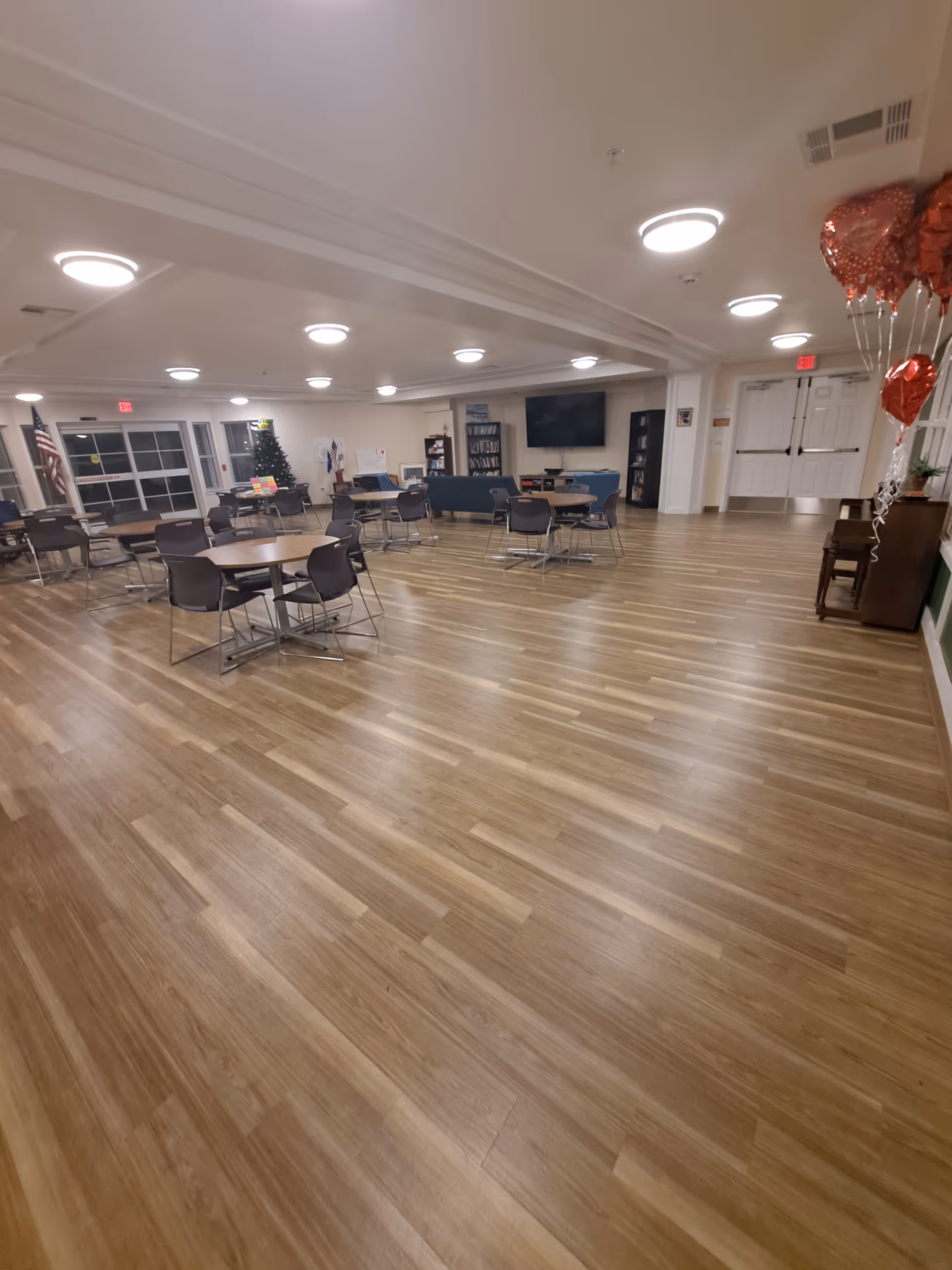 A spacious senior living common area with round tables, chairs, wood floors, and decorations.