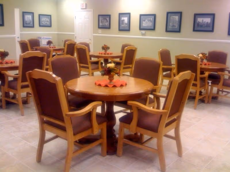 A dining room in a senior living facility with round tables, wooden chairs, and floral centerpieces.