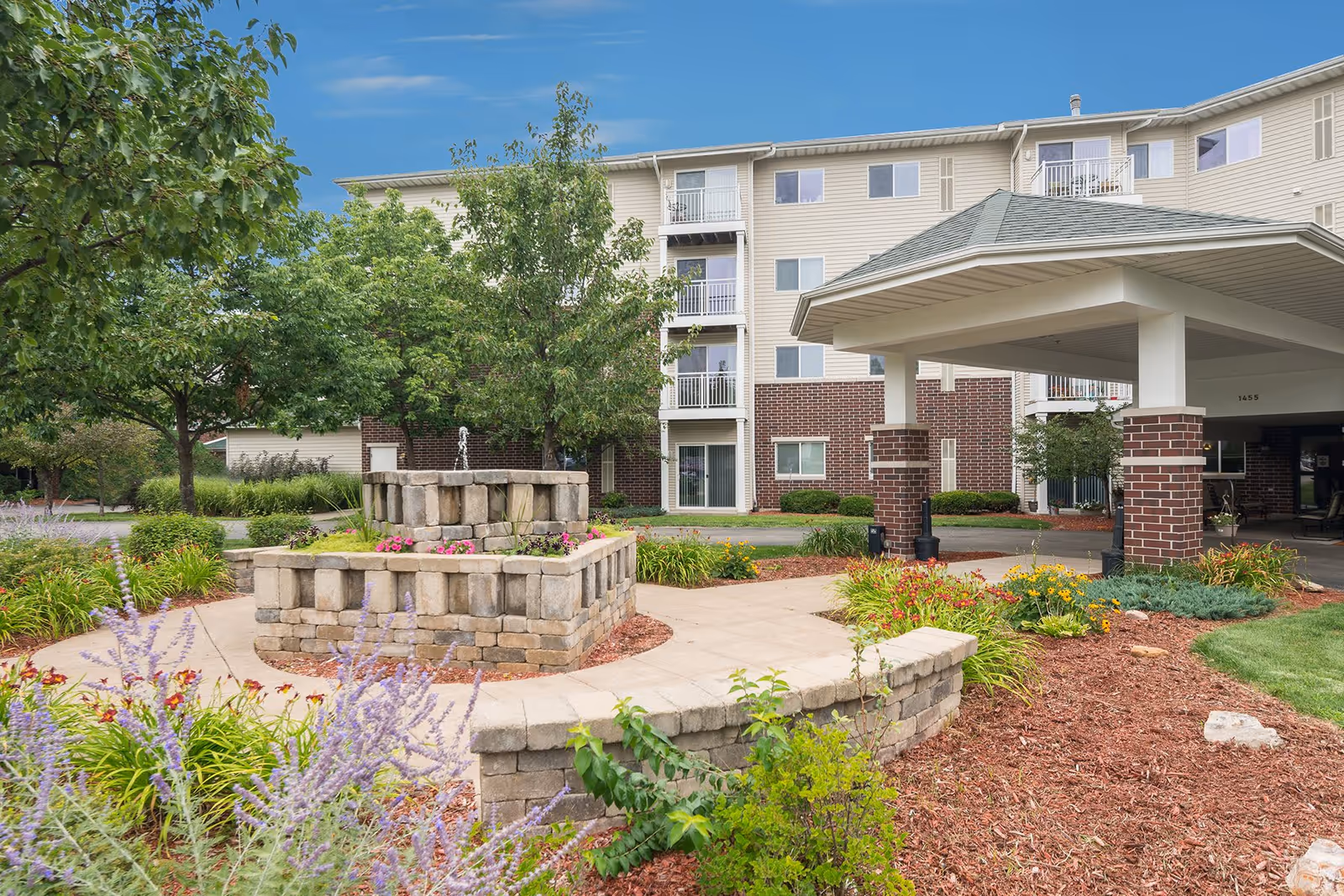 Outdoor view of Park Vista Retirement Living showing a landscaped garden with colorful flowers and greenery, a stone water fountain, and a covered entrance to a multi-story building with balconies and windows under a clear blue sky.