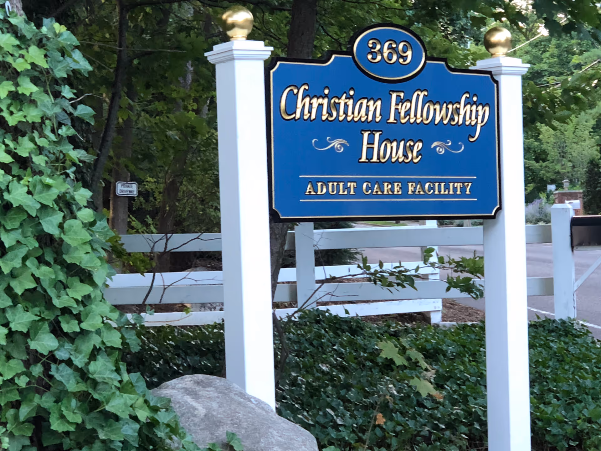 A blue and gold sign reading '369 Christian Fellowship House Adult Care Facility' mounted on white posts with gold finials, surrounded by greenery and a white fence in the background.