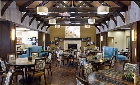 Spacious senior living dining lounge with round wooden tables, teal sofas, and wall art.