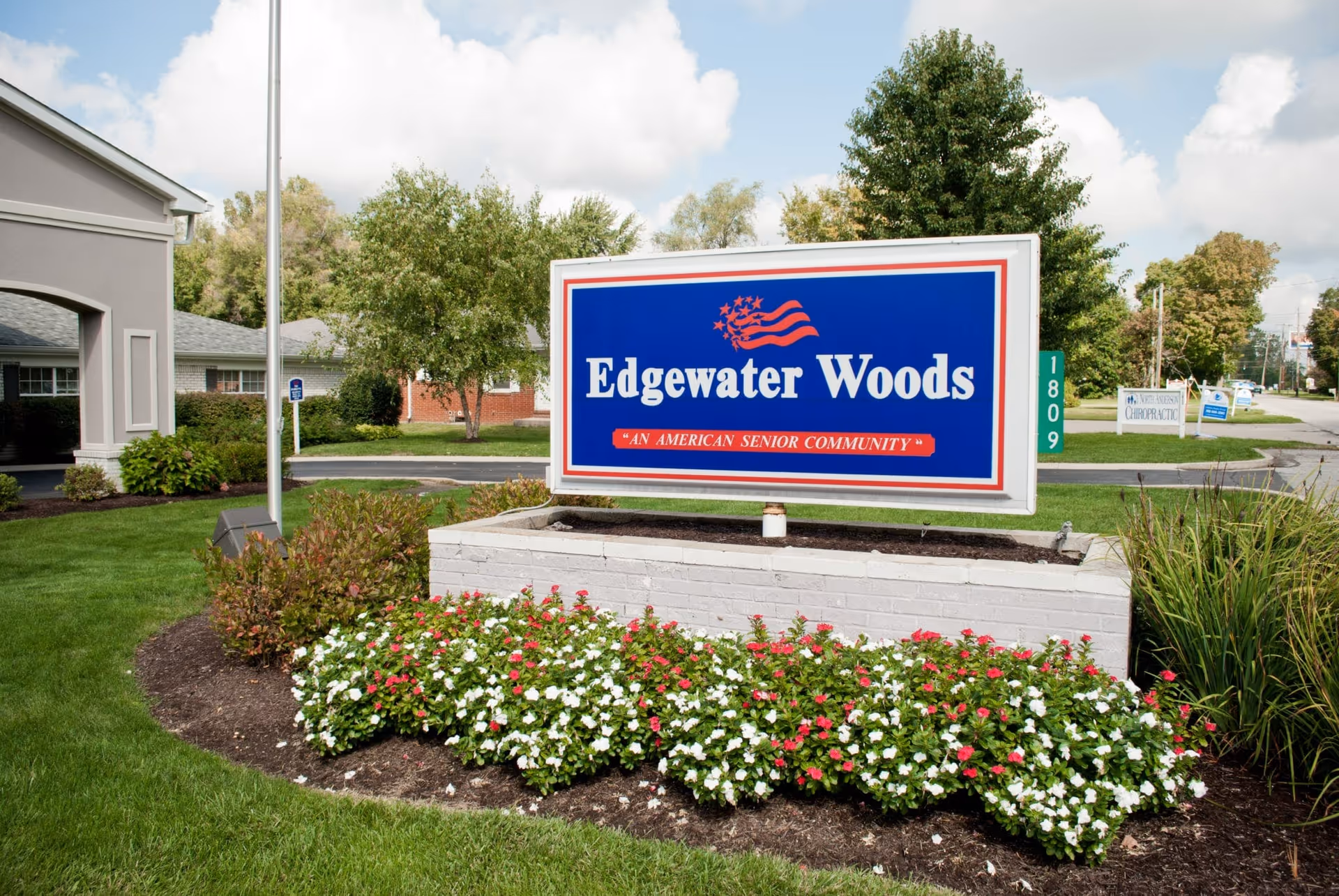 Outdoor view of the Edgewater Woods senior community sign surrounded by a flower bed with red and white flowers, green grass, and trees in the background under a partly cloudy sky.