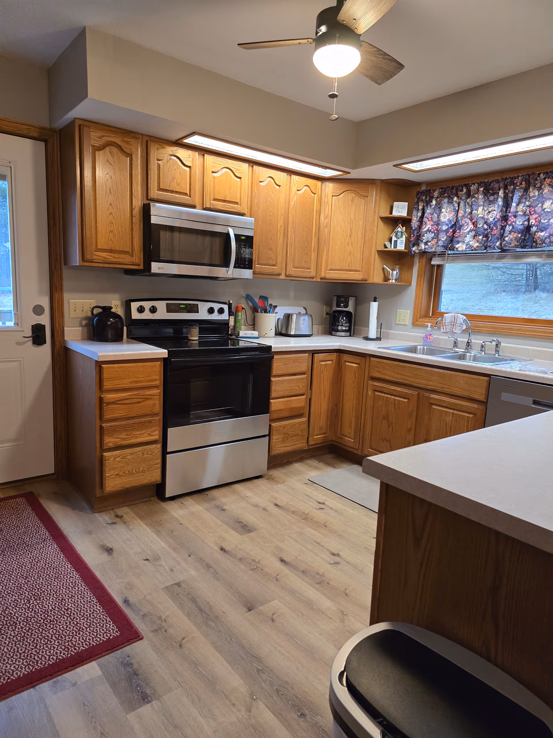 Wooden-cabinet kitchen with stainless stove and microwave, sink, and window with floral valance.