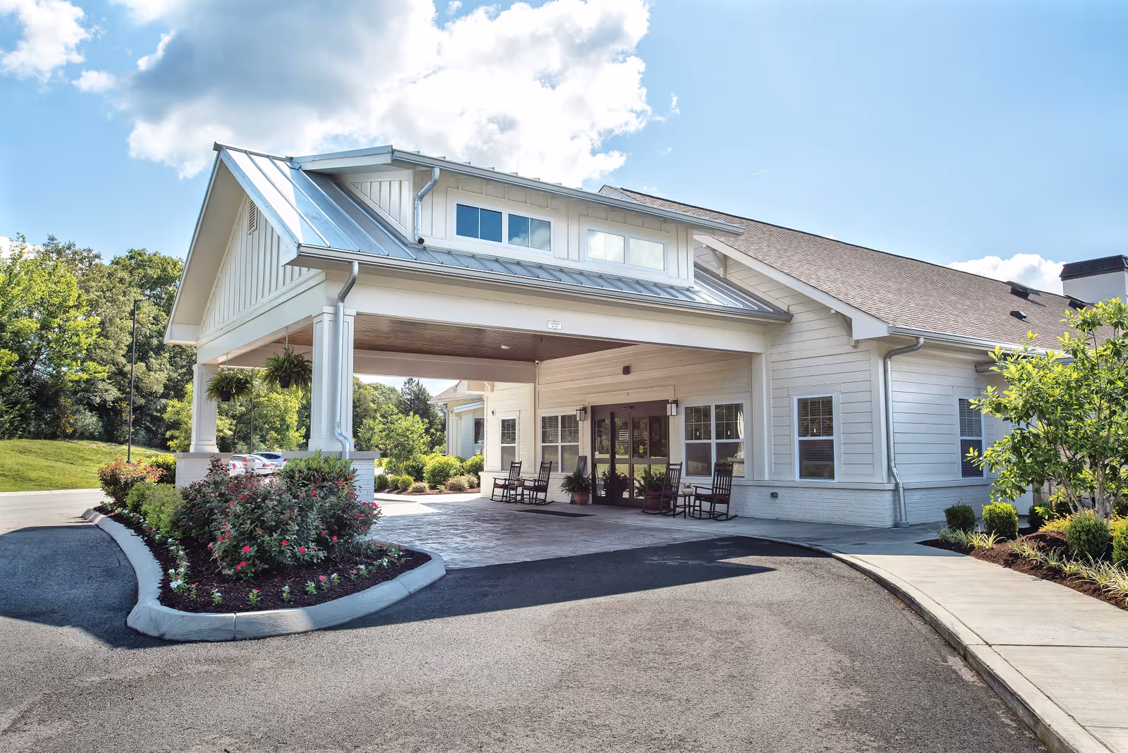 Exterior view of Dominion Senior Living of Athens showing the front entrance with a covered driveway, rocking chairs on the porch, landscaped flower beds, and a clear blue sky with some clouds.