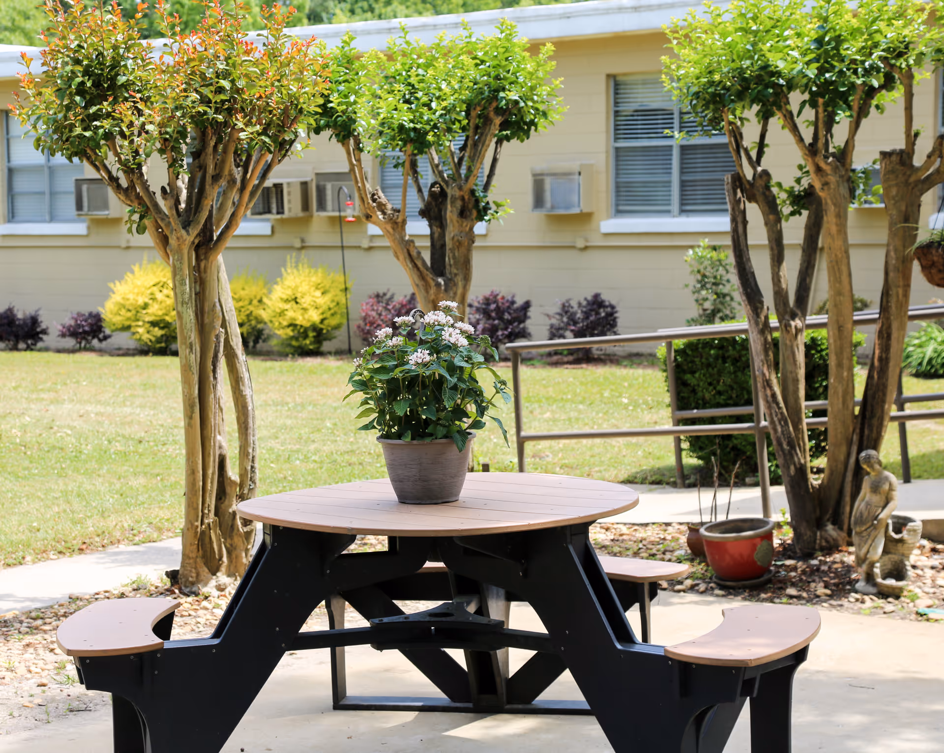 Outdoor patio area with a round picnic table and attached benches. A potted plant with white flowers is placed in the center of the table. Surrounding the area are small trees, shrubs, and a building with windows and air conditioning units.