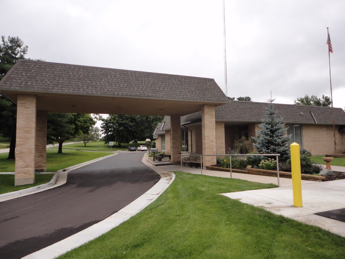 Entrance driveway to a single-story brick building with a covered drop-off area supported by two pillars. There is a paved road leading into the facility, surrounded by green grass and trees. An American flag is visible on a flagpole to the right of the building.