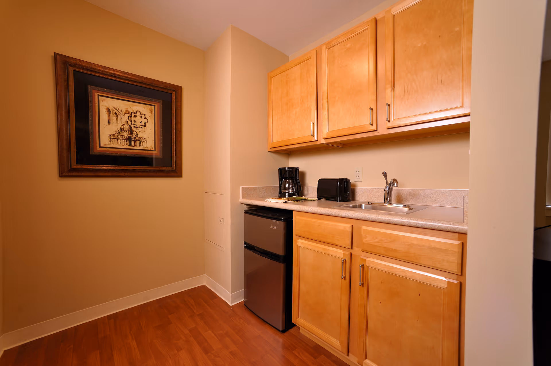 Compact kitchenette with wooden cabinets, sink, mini fridge, toaster, and coffee maker.