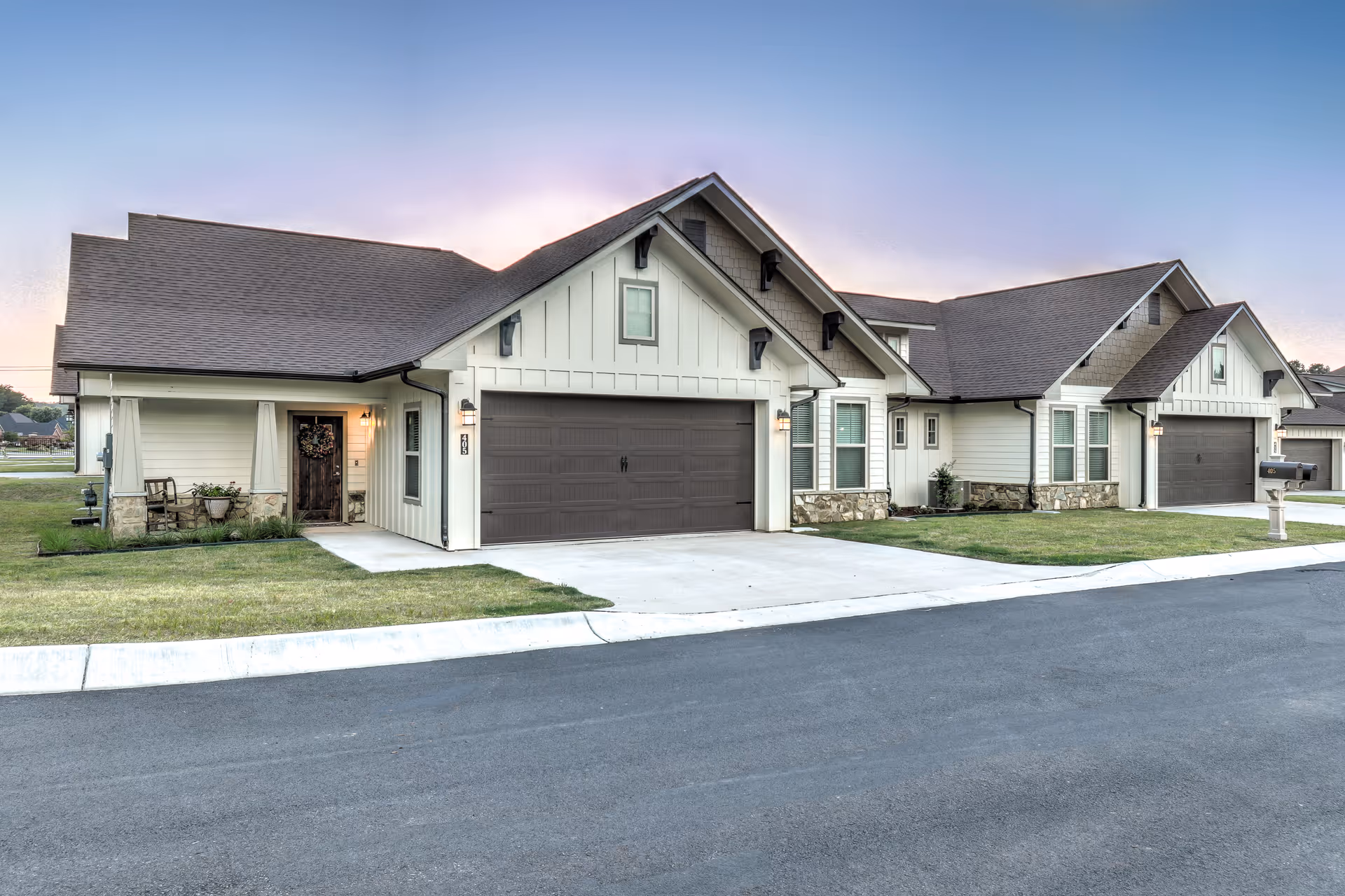 Exterior view of a modern single-story residential building with a large garage door, a small porch with chairs and plants, and a well-maintained lawn. The sky is clear with a soft gradient from blue to light purple, indicating either sunrise or sunset.