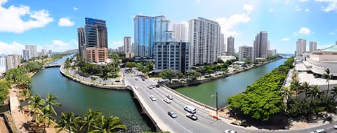 A sunny Waikiki cityscape with a canal, bridge, high-rise buildings, and palm trees.