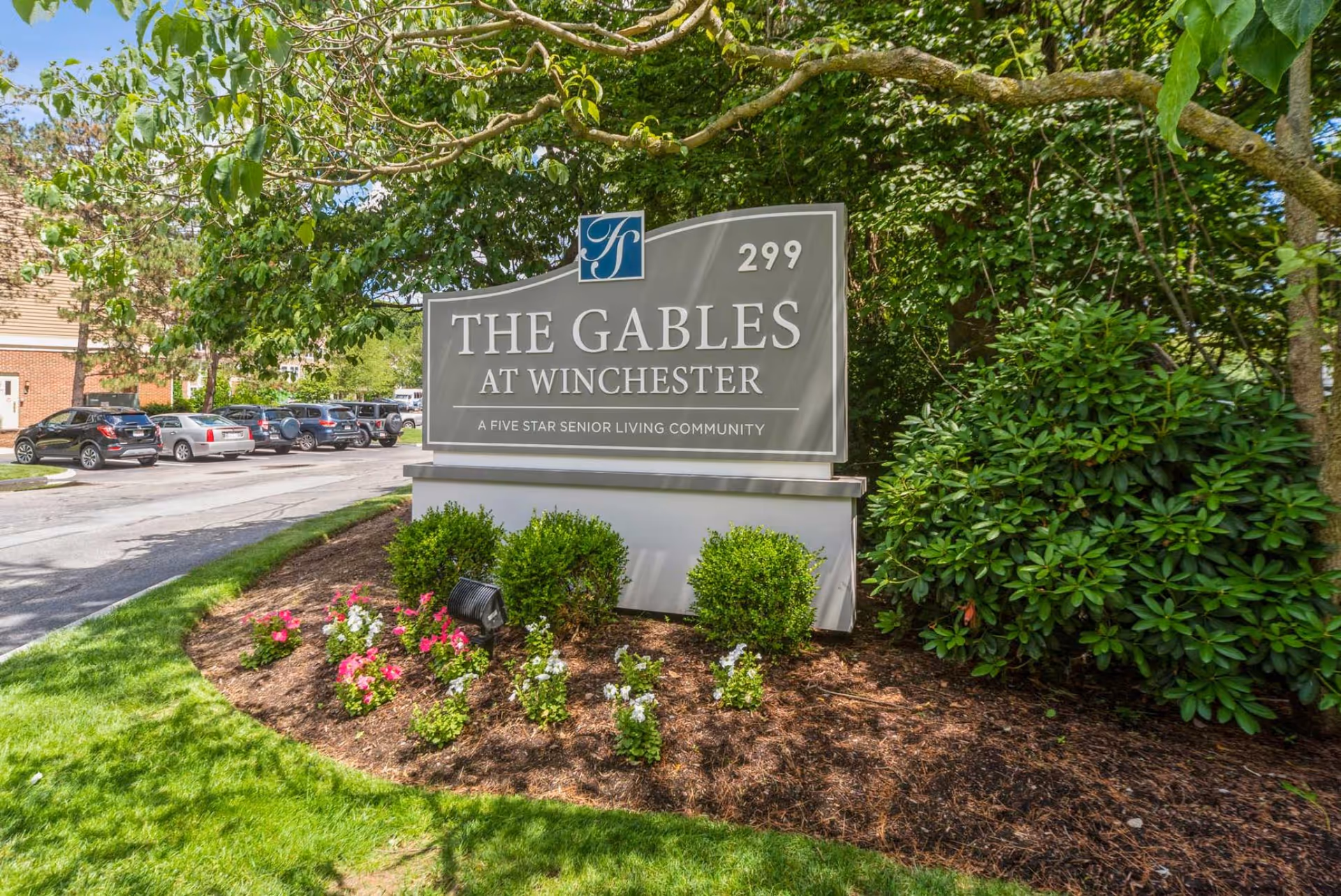 Entrance sign reading 'The Gables at Winchester' surrounded by landscaped flower beds, trees, and parked cars.