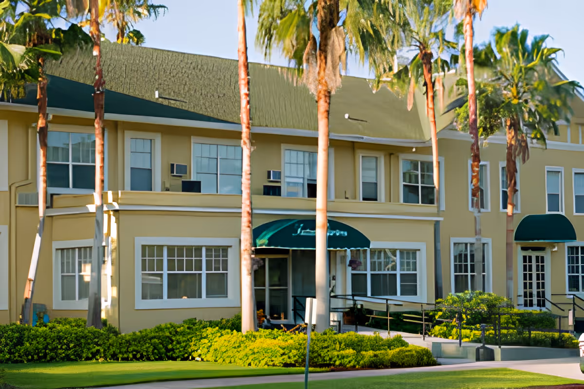 Front exterior of a two-story yellow senior living building with green awnings, palm trees, and landscaped lawn.