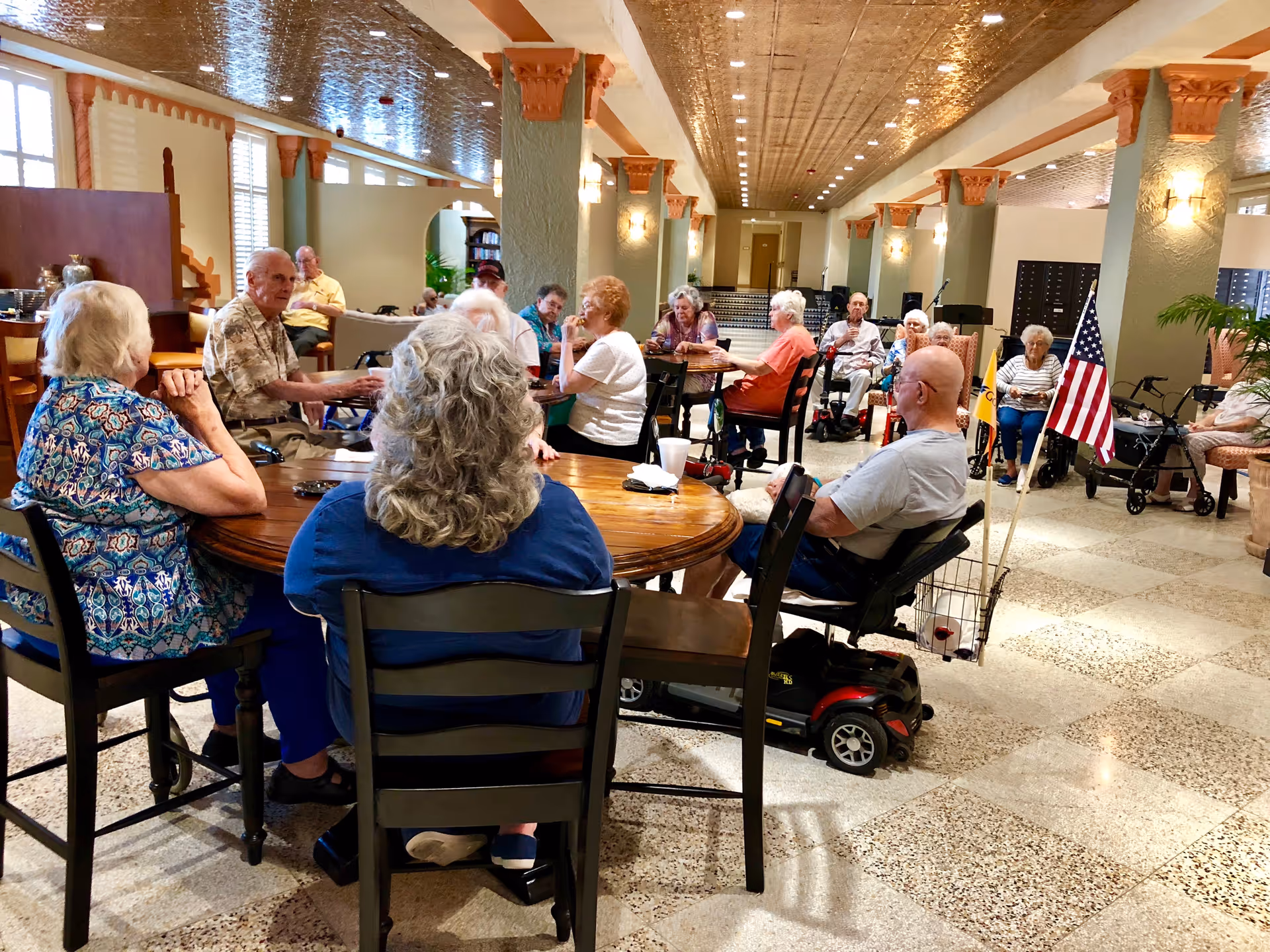 A group of elderly people sitting around tables in a spacious, well-lit common area with decorative columns and a patterned ceiling. Some individuals are seated in wheelchairs or mobility scooters, and the room has a warm, welcoming atmosphere with American flags visible near one person.