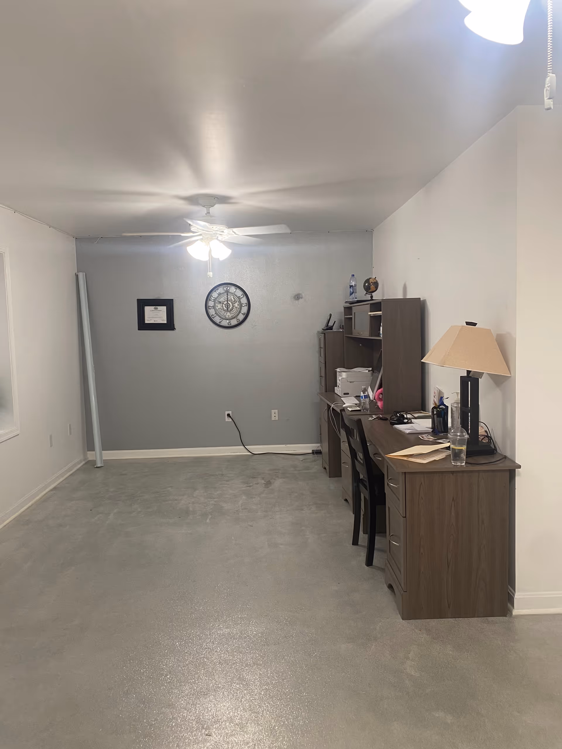 A dim basement office area with a desk, shelves, and a clock on gray walls.
