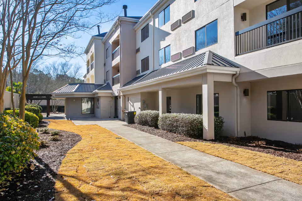 Exterior view of a multi-story senior living building with a covered walkway, concrete path, and landscaped shrubs.