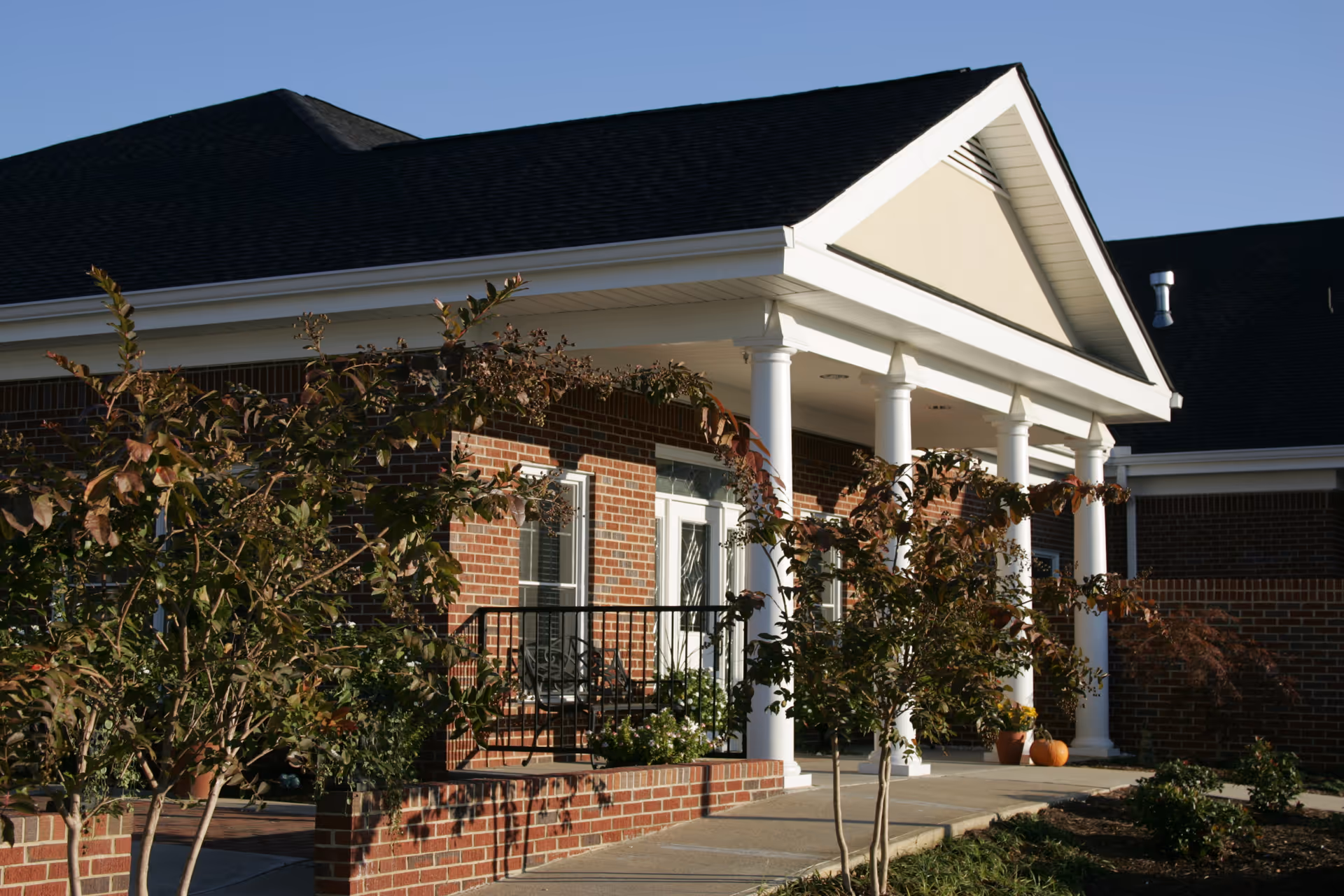 Brick building with a white-columned porch and trees along a paved entryway.