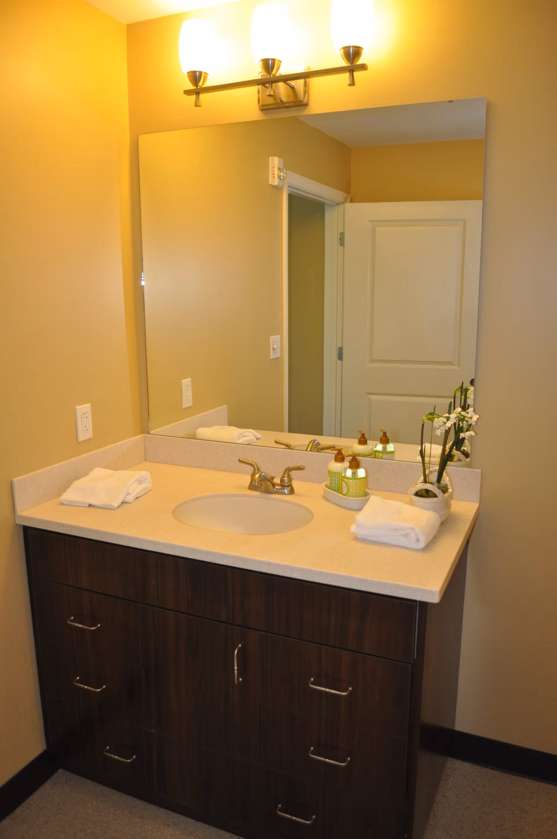 A bathroom vanity with a beige countertop, an oval sink, and a silver faucet. On the countertop are neatly folded white towels, a small potted plant with white flowers, and two bottles of liquid soap or lotion in a small tray. Above the vanity is a large mirror reflecting a partially open white door. A three-light fixture with frosted glass shades is mounted above the mirror. The walls are painted a warm beige color.