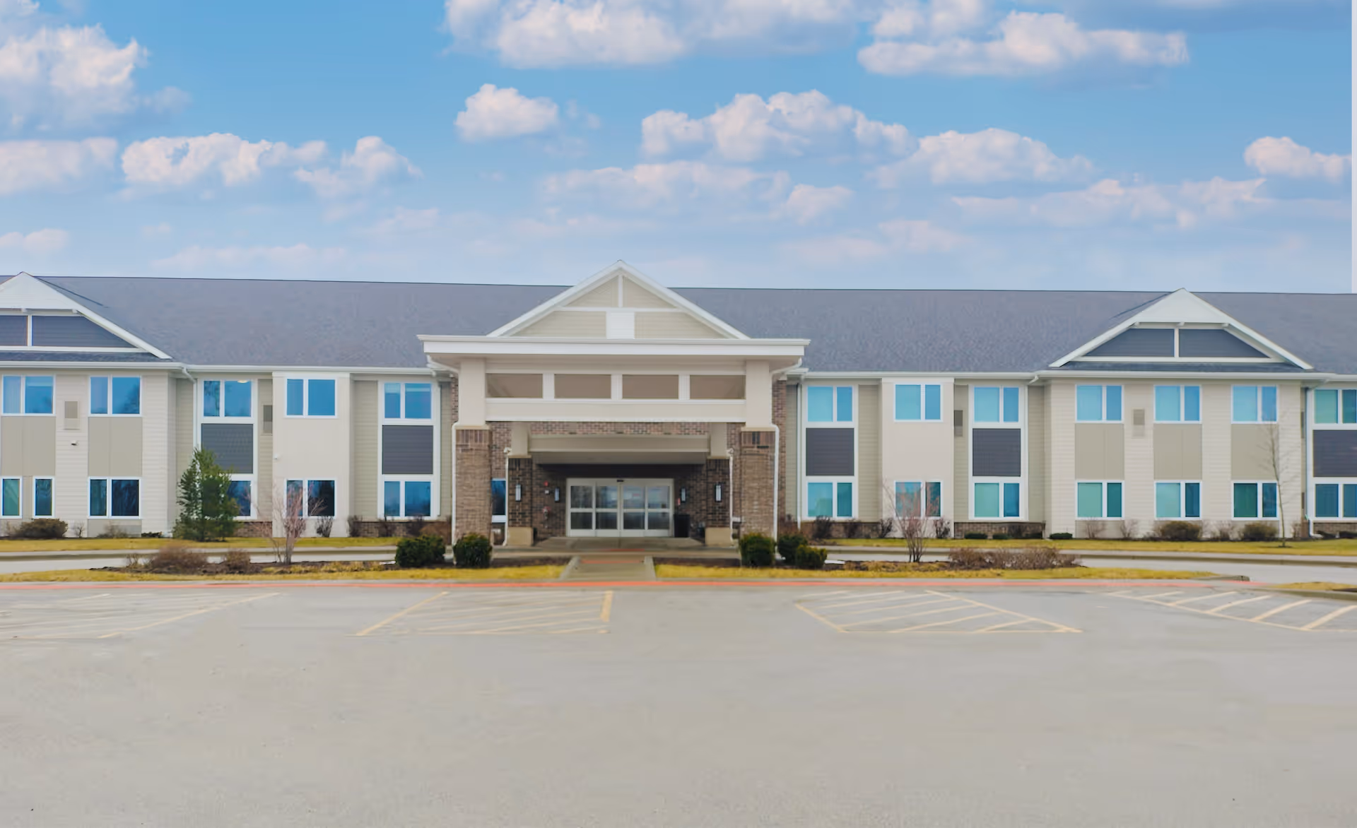 Front exterior view of Ridgeland Senior Living building with a large covered entrance, multiple windows, and a spacious parking lot in front under a partly cloudy sky.