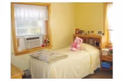 Yellow-walled bedroom with a single bed, window air conditioner, shelf, and a stuffed bear.
