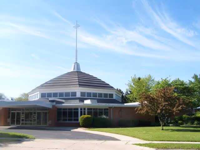 Exterior view of a single-story building with a unique round roof topped by a tall spire. The building is surrounded by green grass, trees, and a clear blue sky with wispy clouds.