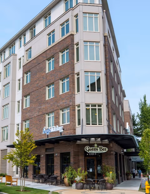 Exterior view of a multi-story brick and beige building with large windows. The building has a sign that reads 'Aegis Living' and a cafe named 'Queen Bee' with outdoor seating and potted plants. There are small trees and a bench on the sidewalk in front of the building.