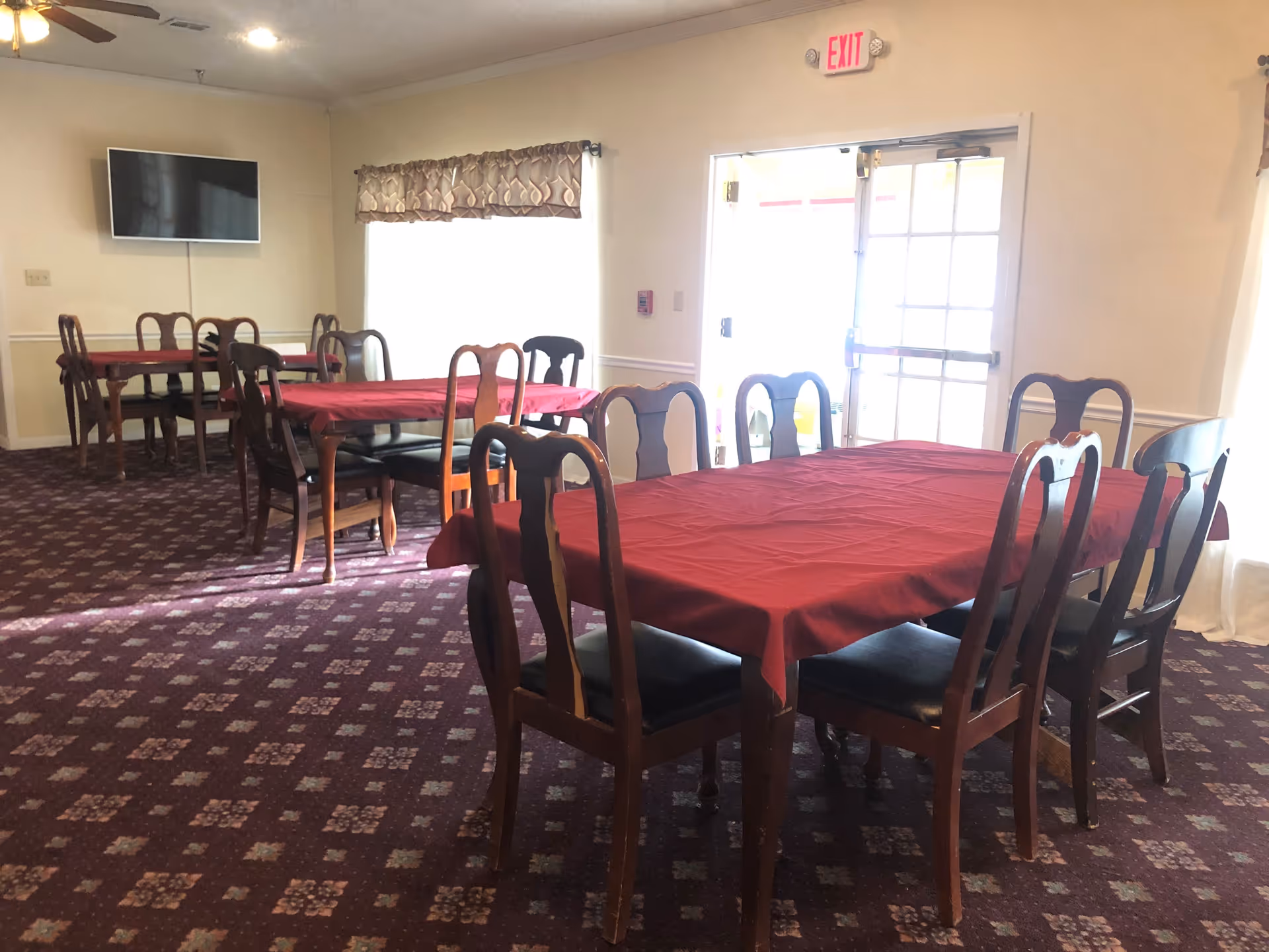 Interior of a dining room with multiple wooden tables covered with red tablecloths and surrounded by wooden chairs with black cushions. The room has patterned carpet flooring, a wall-mounted flat screen TV, a ceiling fan, and large windows with valance curtains. A glass door with an exit sign above it is visible on the right side.