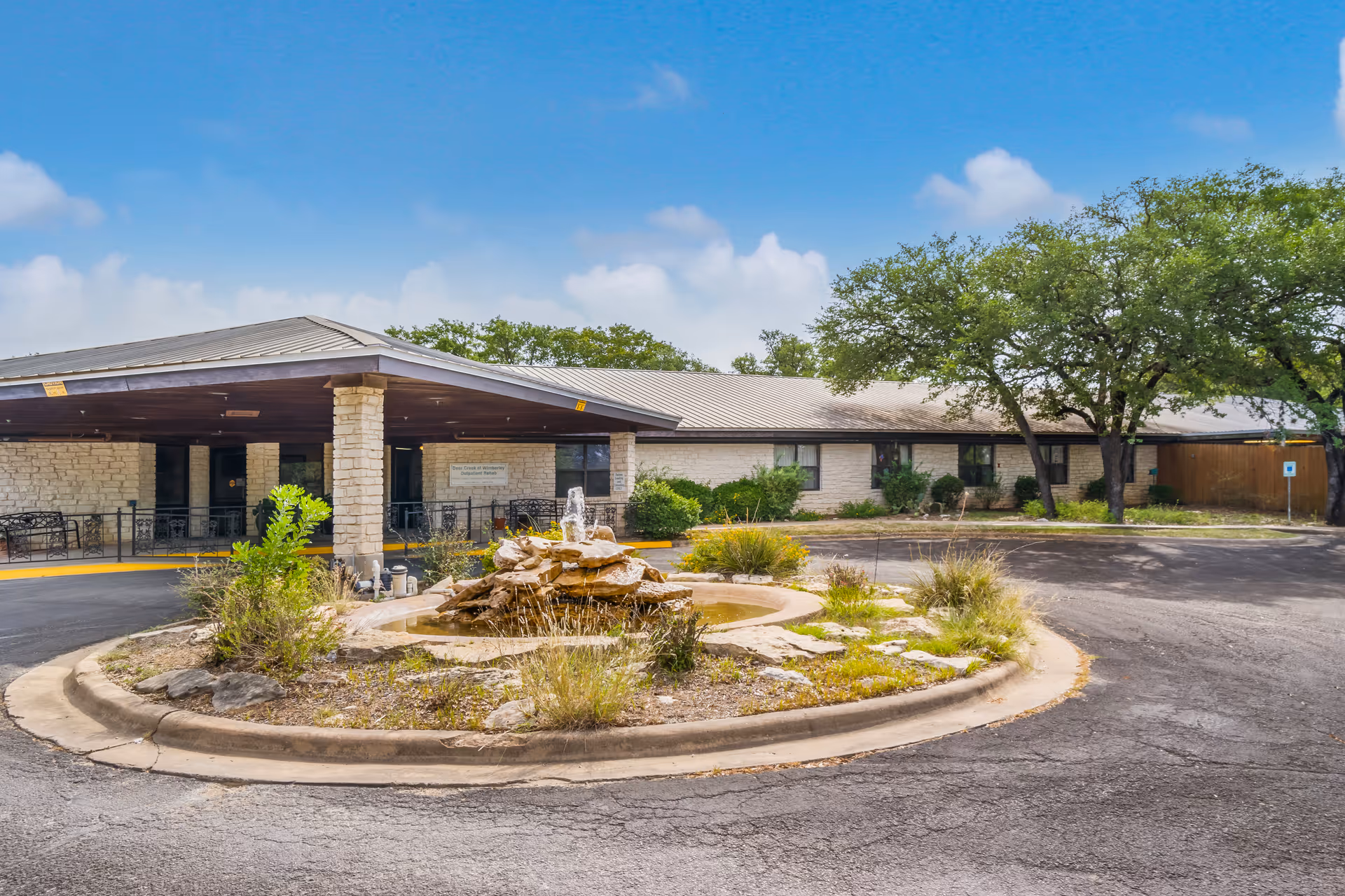 Exterior view of Deer Creek Nursing and Rehab facility showing a single-story building with a covered entrance supported by stone pillars. In front of the entrance is a circular driveway with a small rock fountain and landscaping. Trees and shrubs surround the building under a partly cloudy blue sky.