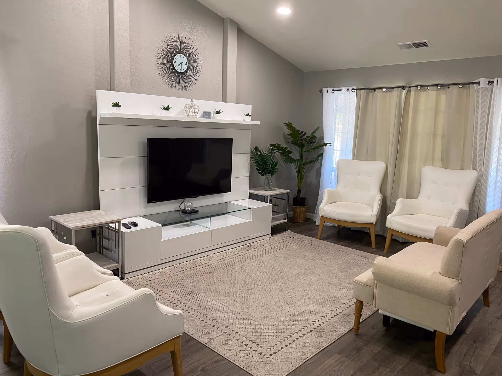 A modern living room with a large flat-screen TV mounted on a white entertainment unit. The room features four white upholstered chairs and a beige upholstered bench arranged around a large beige area rug. There are two side tables, a decorative clock on the wall above the TV, and potted plants near the window with beige curtains.